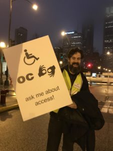 Smiling man with large sign featuring large acessibility symbols and the text "ask me about access!"