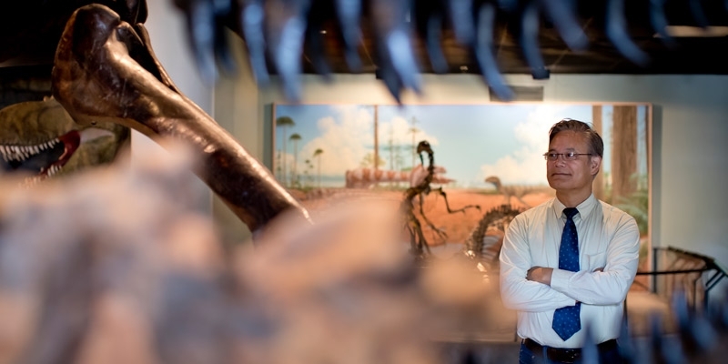 Man standing with folded arms in an exhibit hall with dinosaur skeleton in foreground