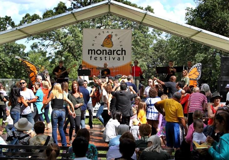photograph of the festival scene, a several people in front of a stage, musicians on the stage along with two people wearing orange and black costume butterfly wings, and a large sign hanging over the stage with the words "Monarch Festival". Trees and blue sky in the background.