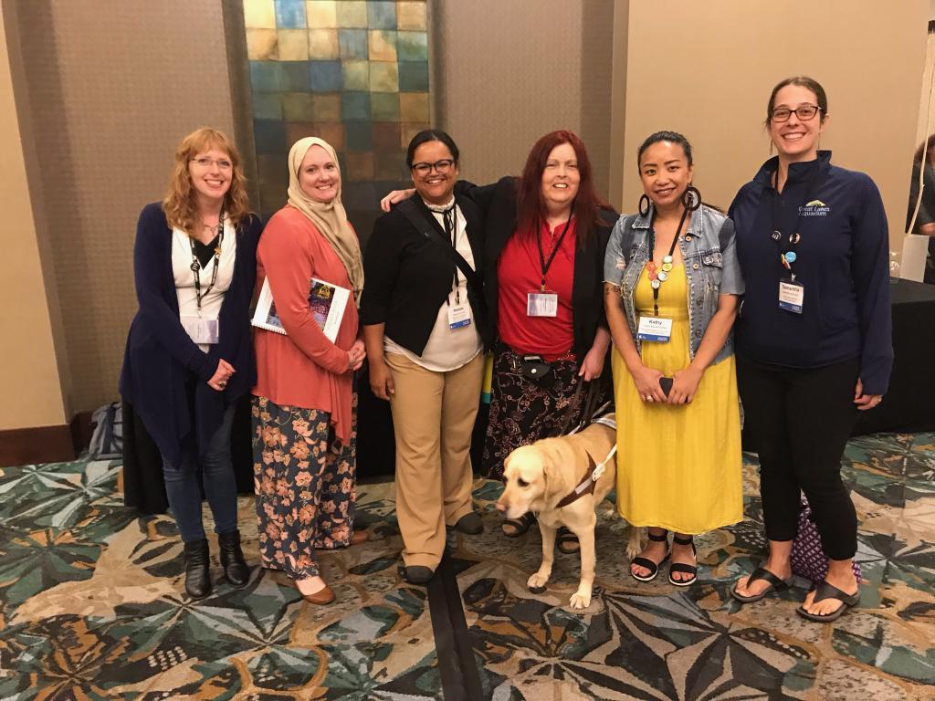 Six smiling feminine people stand in a row with a Yellow Labrador in front. From left to right: Emma Allen, Bell Museum; Jenea Rewertz-TArgui, Ordway; Natalie Kennedy-Schuck, MSAB; Sherry Shireck, Arts Access for All; Millie, Yellow Labrador; Kathy Mouacheupao, MRAC; and Samantha Smingler, Great Lakes Aquarium.